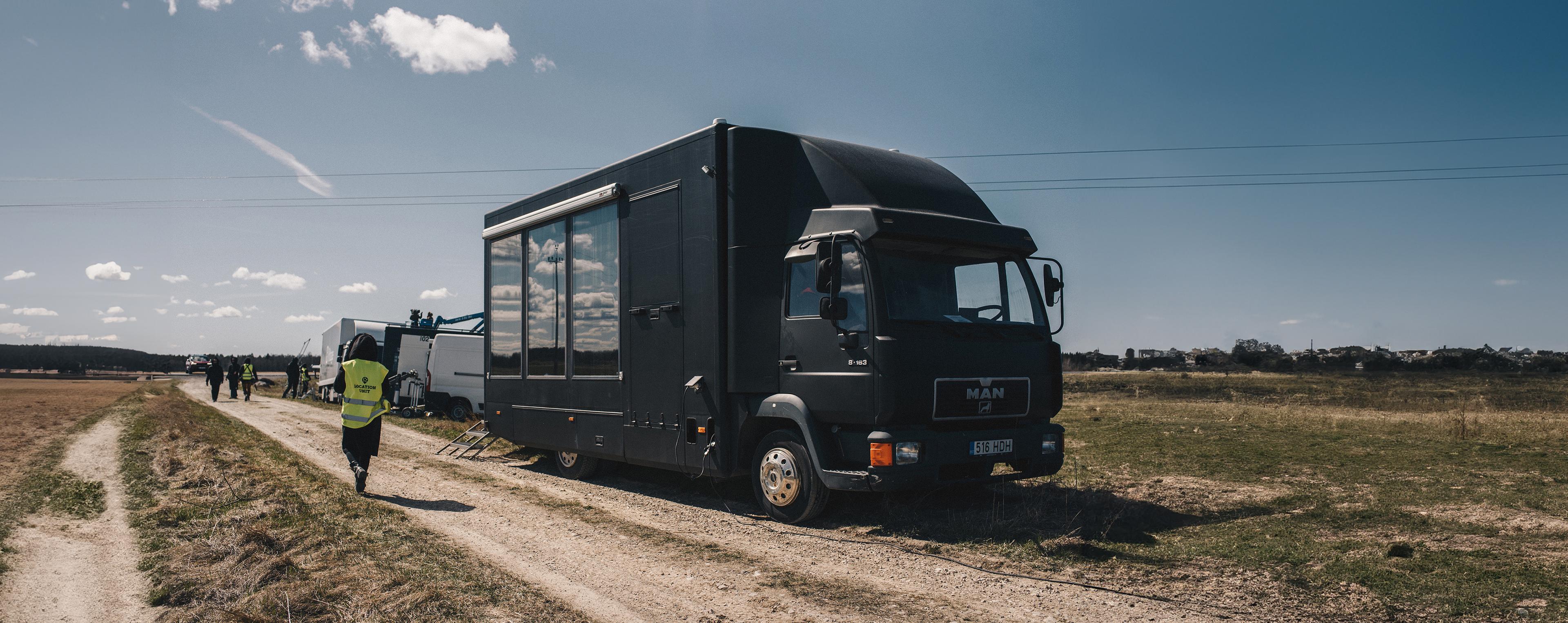 Film crew working with cars and equipment on the side of a dirt road that goes through a field.